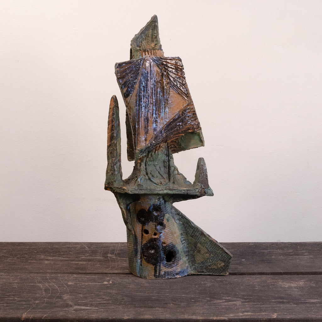 An abstract sculptural art piece with a textured, weathered finish in earthy tones of brown, green, and blue, displayed on a dark wooden surface against a plain off-white wall.