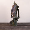 An abstract sculptural art piece with a textured, weathered finish in dark green, brown, and black, displayed on a rustic wooden surface against a plain off-white background.