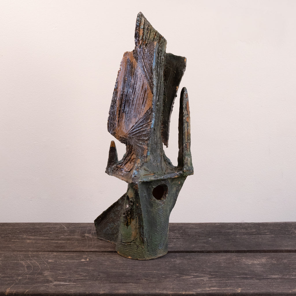 An abstract sculptural art piece with a textured, weathered finish in dark green, brown, and black, displayed on a rustic wooden surface against a plain off-white background.