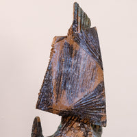 A close-up of a textured ceramic sculpture featuring a stylized fan or sail-like geometric form in earthy brown, bronze, and blue glazes against a plain light beige background.