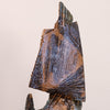 A close-up of a textured ceramic sculpture featuring a stylized fan or sail-like geometric form in earthy brown, bronze, and blue glazes against a plain light beige background.