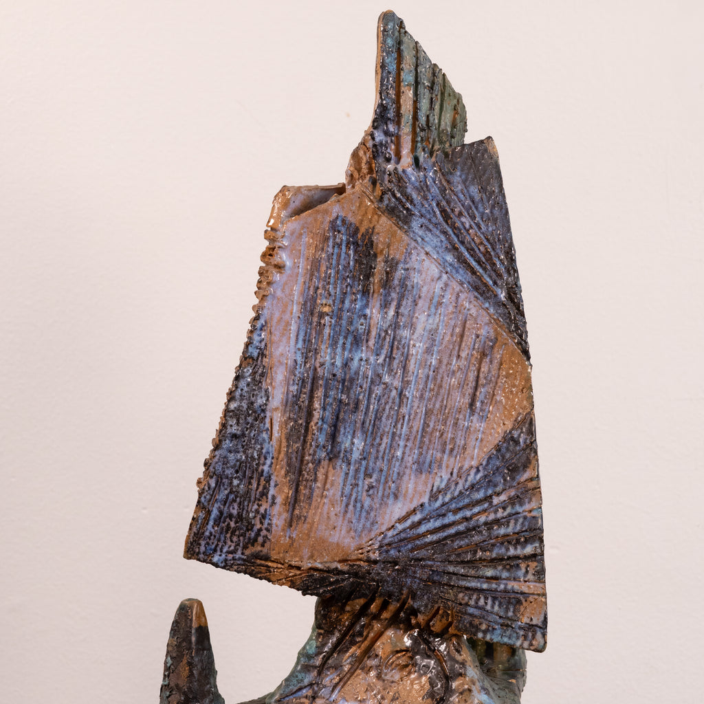 A close-up of a textured ceramic sculpture featuring a stylized fan or sail-like geometric form in earthy brown, bronze, and blue glazes against a plain light beige background.