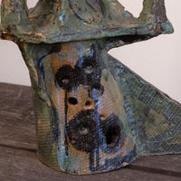 A close-up of a textured ceramic sculpture with a distressed, weathered finish in earthy green, blue, brown, and black glazes, featuring a series of circular holes on its front.