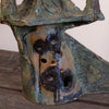 A close-up of a textured ceramic sculpture with a distressed, weathered finish in earthy green, blue, brown, and black glazes, featuring a series of circular holes on its front.