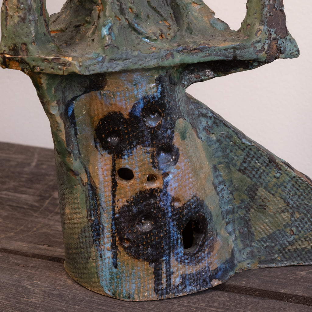 A close-up of a textured ceramic sculpture with a distressed, weathered finish in earthy green, blue, brown, and black glazes, featuring a series of circular holes on its front.