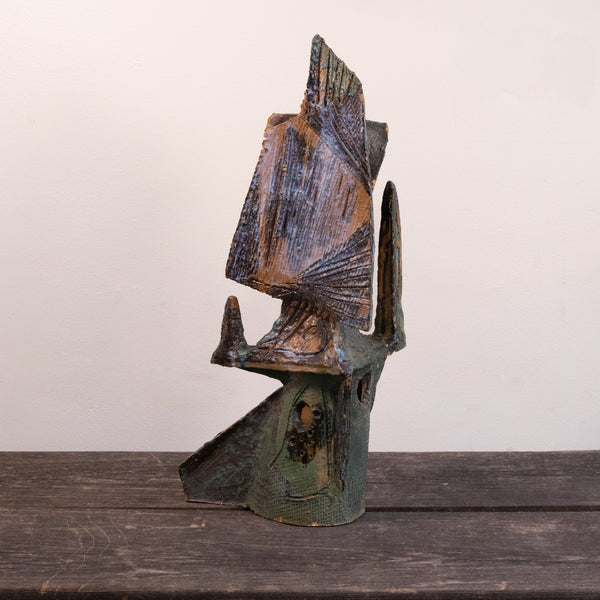 A rustic, handcrafted ceramic sculpture of an abstract figure, featuring weathered brown and green patina textures, displayed on a dark wooden surface against a plain off-white wall.