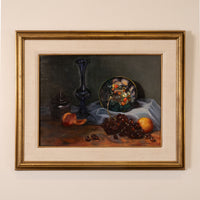 A framed still life painting on a beige wall, featuring a dark blue glass vase, a glass bowl of fruit, grapes, and peaches on a draped cloth.