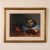 A framed still life painting on a beige wall, featuring a dark blue glass vase, a glass bowl of fruit, grapes, and peaches on a draped cloth.