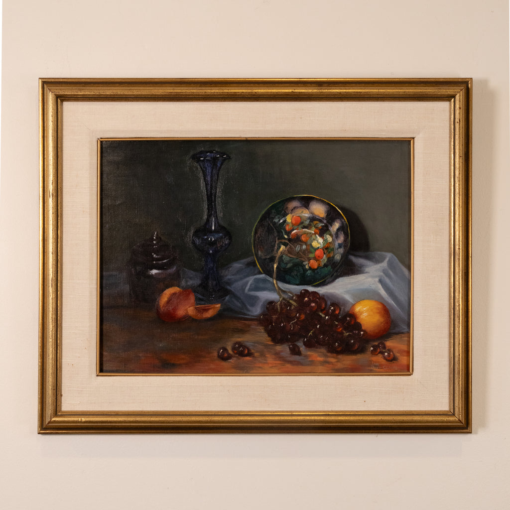 A framed still life painting on a beige wall, featuring a dark blue glass vase, a glass bowl of fruit, grapes, and peaches on a draped cloth.