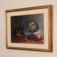 A framed still life oil painting featuring a dark blue glass vase, a plate of fruit, grapes, and an orange on a draped cloth, presented in a wide gold frame with a cream mat against a neutral wall.