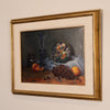 A framed still life oil painting featuring a dark blue glass vase, a plate of fruit, grapes, and an orange on a draped cloth, presented in a wide gold frame with a cream mat against a neutral wall.