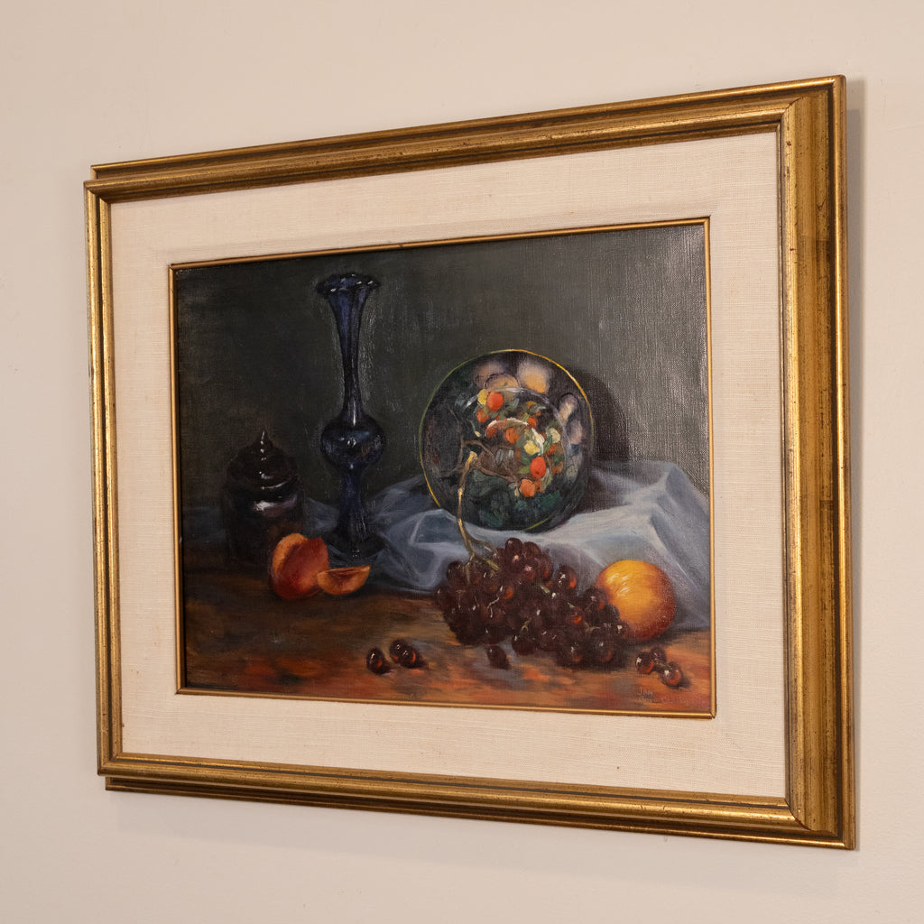 A framed still life oil painting featuring a dark blue glass vase, a plate of fruit, grapes, and an orange on a draped cloth, presented in a wide gold frame with a cream mat against a neutral wall.