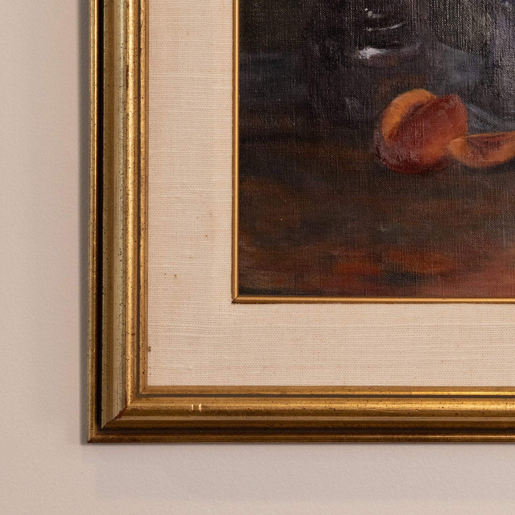 Close-up of a framed still life painting showing part of a gold ornate frame, cream mat board, and dark brown painted canvas with orange fruit details.