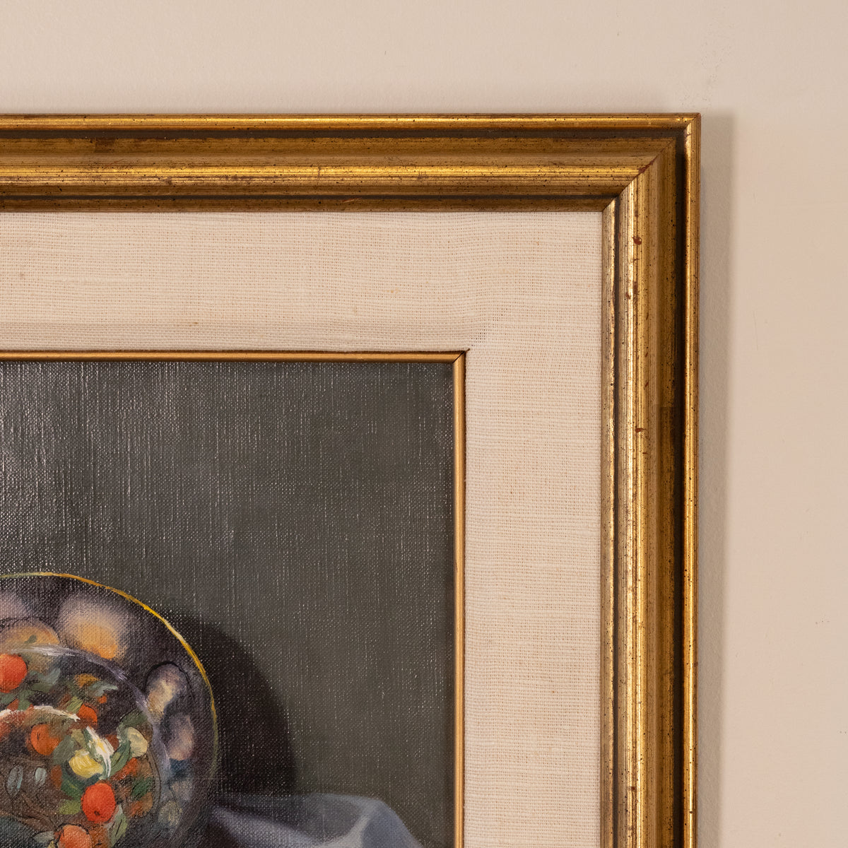 Close-up of a framed painting showing part of a still life scene with a colorful bowl and dark gray background, bordered by a wide beige mat and ornate gold frame against a light wall.