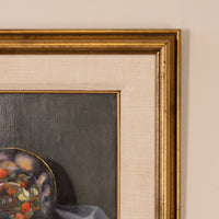 Close-up of a framed painting showing part of a still life scene with a colorful bowl and dark gray background, bordered by a wide beige mat and ornate gold frame against a light wall.