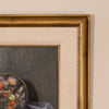 Close-up of a framed painting showing part of a still life scene with a colorful bowl and dark gray background, bordered by a wide beige mat and ornate gold frame against a light wall.