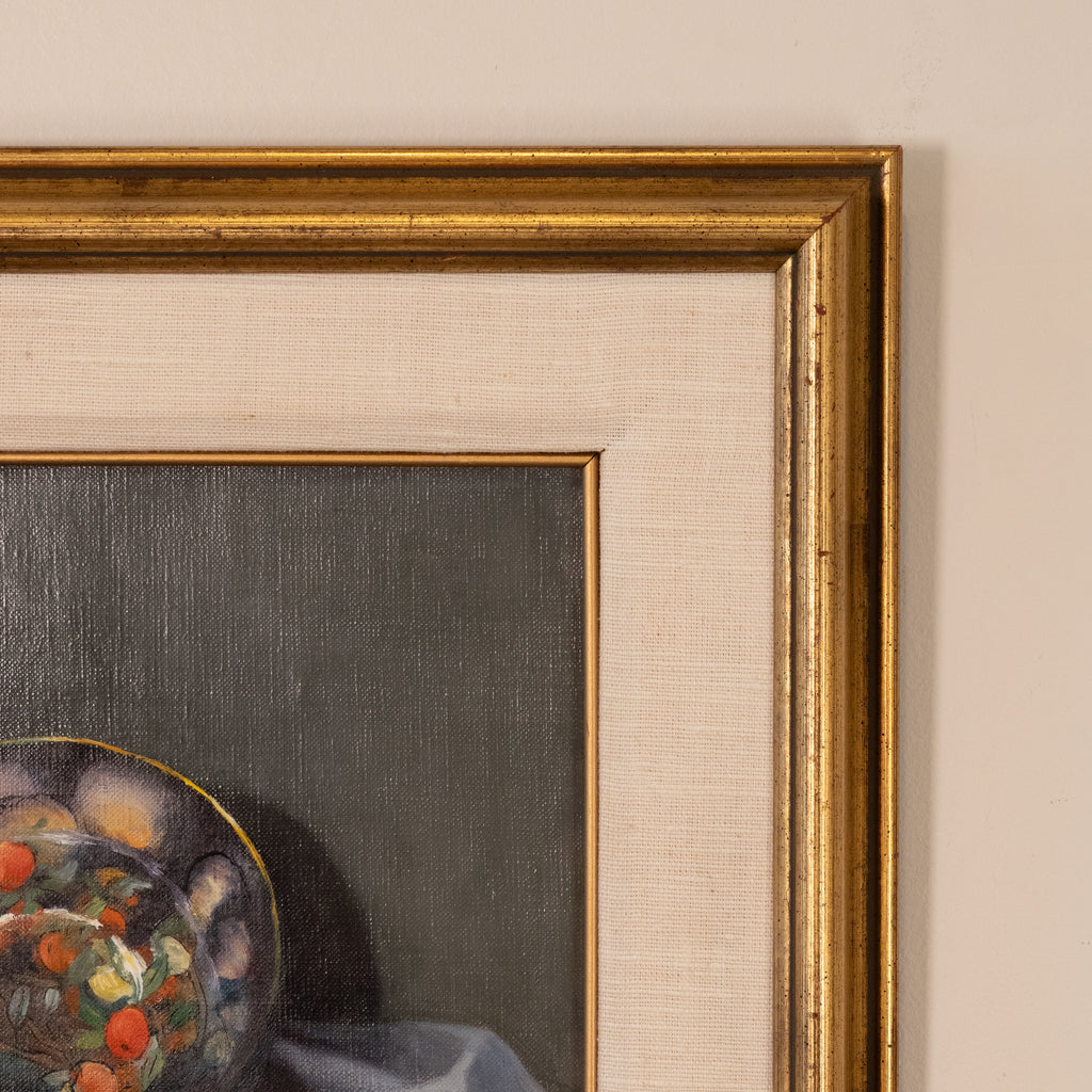 Close-up of a framed painting showing part of a still life scene with a colorful bowl and dark gray background, bordered by a wide beige mat and ornate gold frame against a light wall.