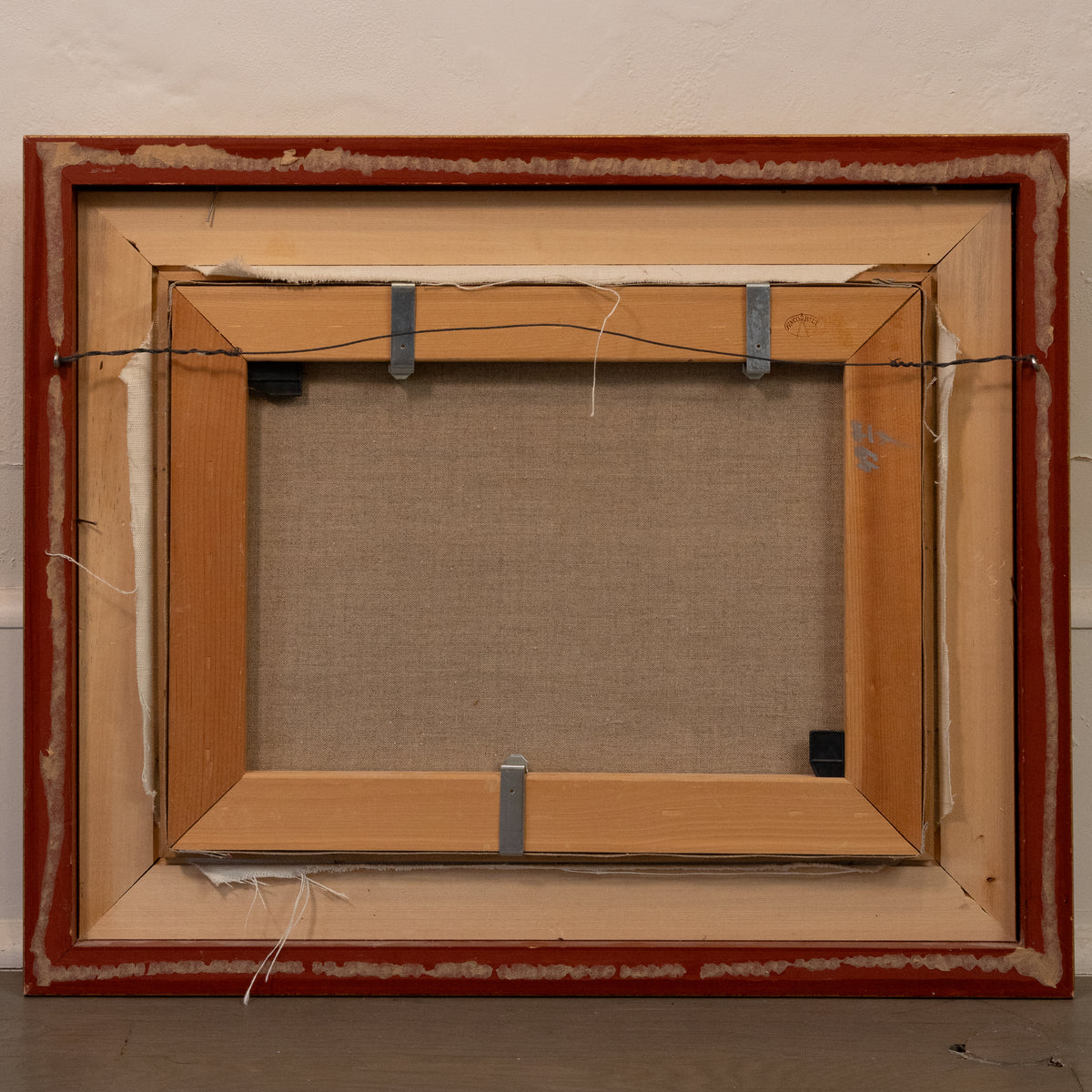 The back view of a framed canvas painting, showing the wooden frame, canvas fabric backing, and hanging hardware against a textured off-white wall.