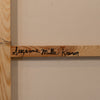 A close-up of a wooden art canvas or print frame, showing a light natural wood bar with the name 'Jazmine Milla Kramer' written in black script, mounted against a beige wall.