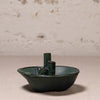A small handmade ceramic vessel with a dark green glaze and two upright cylindrical handles, photographed against a textured off-white wall and neutral surface.