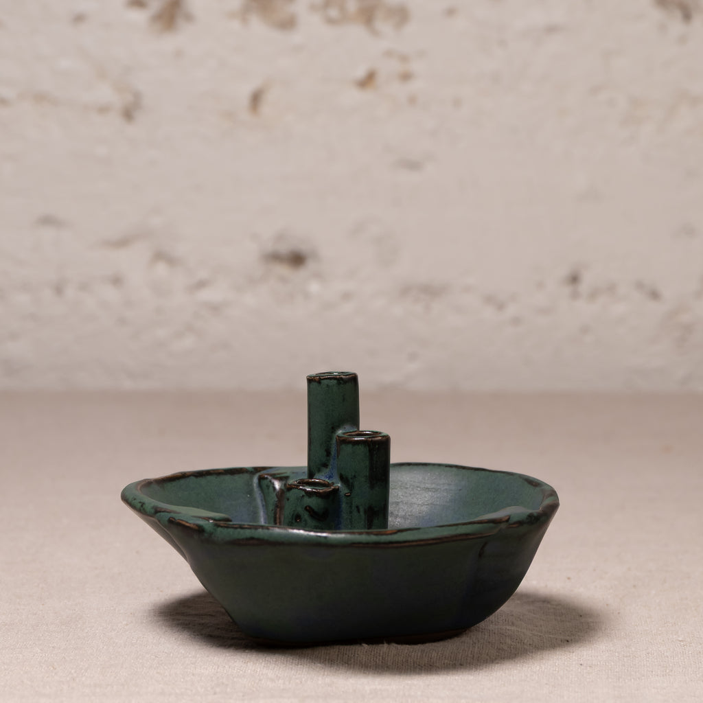 A small handmade ceramic vessel with a dark green glaze and two upright cylindrical handles, photographed against a textured off-white wall and neutral surface.