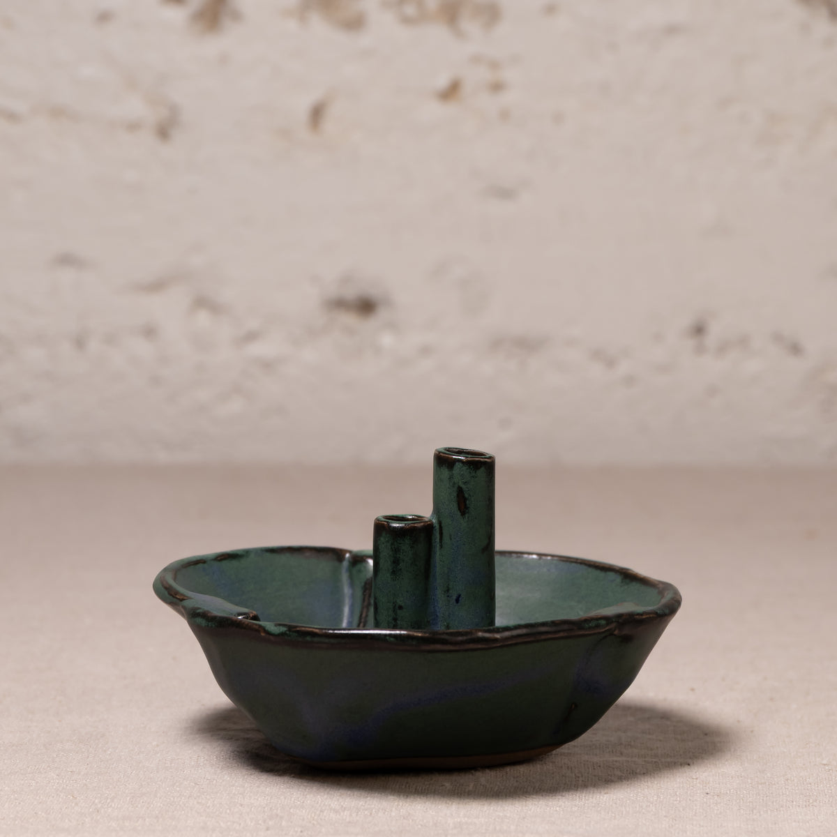 A small handmade ceramic dish with two vertical cylindrical handles, featuring a dark green and blue-green glazed finish, set against a textured light beige background.