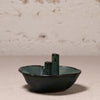 A small handmade ceramic dish with two vertical cylindrical handles, featuring a dark green and blue-green glazed finish, set against a textured light beige background.
