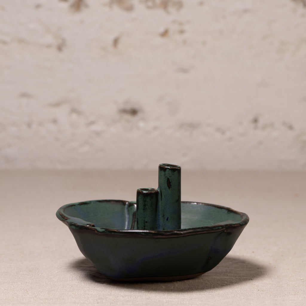 A small handmade ceramic dish with two vertical cylindrical handles, featuring a dark green and blue-green glazed finish, set against a textured light beige background.