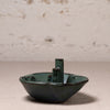 A small ceramic boat-shaped dish or candle holder with a glossy dark green glaze and a central cylindrical stem, photographed on a neutral surface against a textured off-white wall.