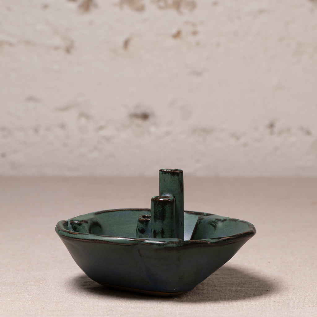 A small ceramic boat-shaped dish or candle holder with a glossy dark green glaze and a central cylindrical stem, photographed on a neutral surface against a textured off-white wall.