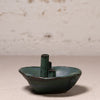A small handmade ceramic bowl with a central cylindrical stem, glazed in a dark teal-green color with a rustic, distressed finish, set against a neutral textured background.