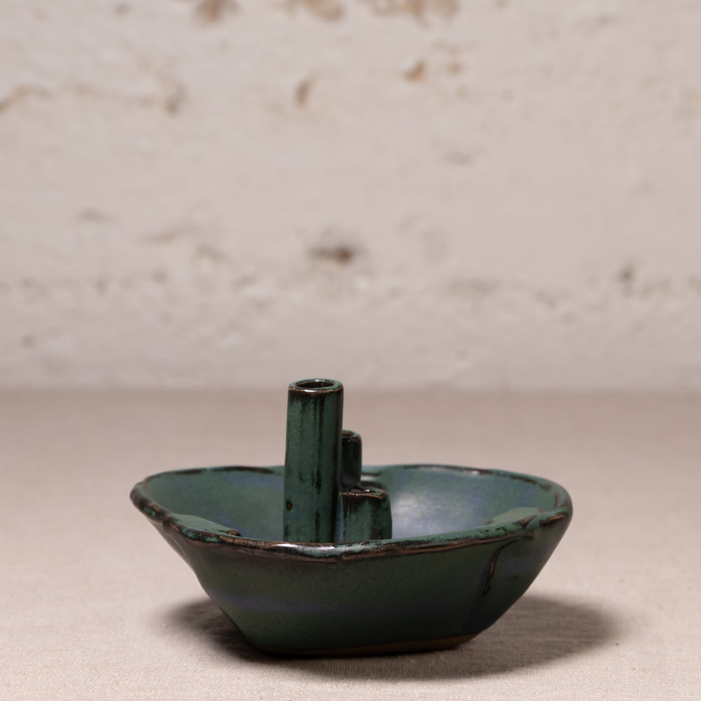 A small handmade ceramic bowl with a central cylindrical stem, glazed in a dark teal-green color with a rustic, distressed finish, set against a neutral textured background.