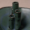 A close-up of a handmade ceramic vase with three vertical cylindrical necks, featuring a dark green and blue-green glaze with brown clay showing around the rims.