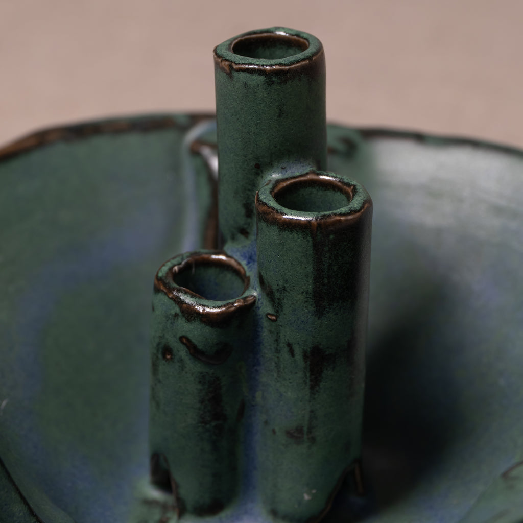 A close-up of a handmade ceramic vase with three vertical cylindrical necks, featuring a dark green and blue-green glaze with brown clay showing around the rims.