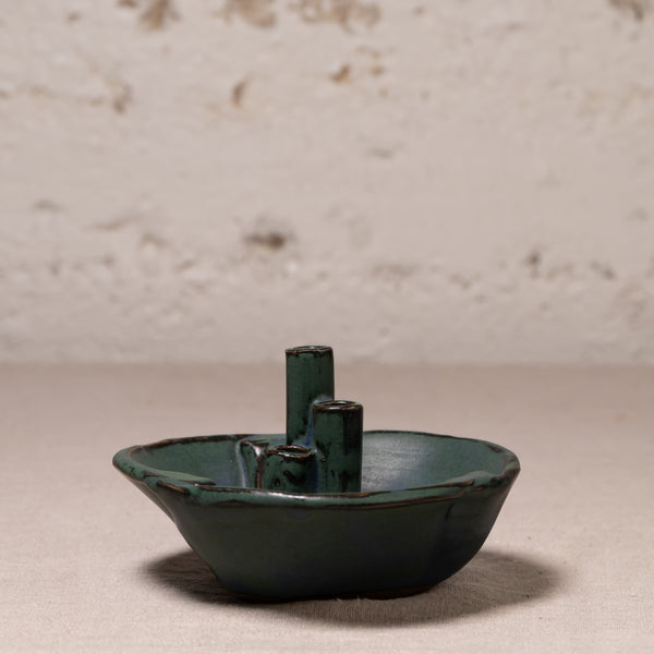 A small handmade ceramic vessel with a dark green glaze and two upright cylindrical handles, photographed against a textured off-white wall and neutral surface.