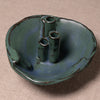 A handmade ceramic dish with three cylindrical openings, featuring a textured glaze in shades of teal, blue, and green with dark brown crackled edges, set against a neutral beige fabric background.