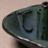 Close-up of a handmade ceramic vessel, likely a candle holder or small dish, featuring a textured glaze in deep teal green with dark brown earthy accents, set against a neutral beige fabric background.