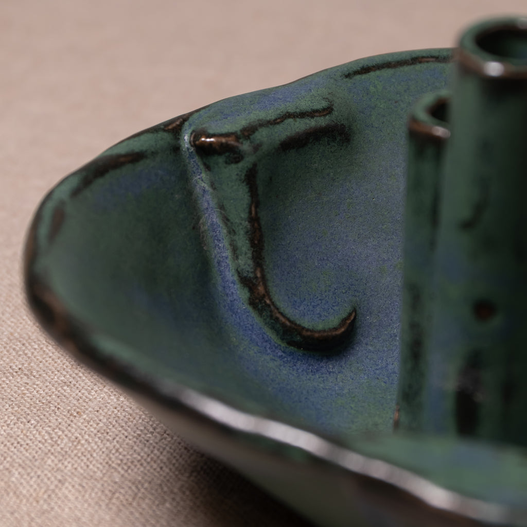 Close-up of a handmade ceramic vessel, likely a candle holder or small dish, featuring a textured glaze in deep teal green with dark brown earthy accents, set against a neutral beige fabric background.