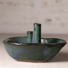 A handmade ceramic candle holder shaped like a small boat, featuring a rounded base, a central pillar, and two smaller side pillars, all glazed in a textured teal green with dark brown speckles and a subtle blue sheen, set against a neutral, softly blurred background.