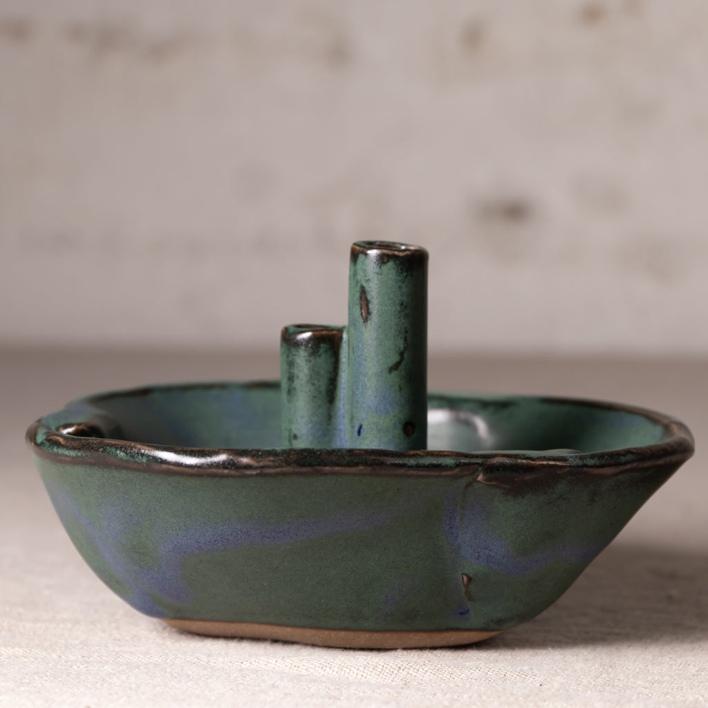 A handmade ceramic candle holder shaped like a small boat, featuring a rounded base, a central pillar, and two smaller side pillars, all glazed in a textured teal green with dark brown speckles and a subtle blue sheen, set against a neutral, softly blurred background.