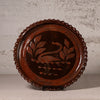 A round dark brown wooden serving tray with a scalloped edge, featuring an inlaid swan and leaf design in the center, displayed on a stand against a textured light gray wall.