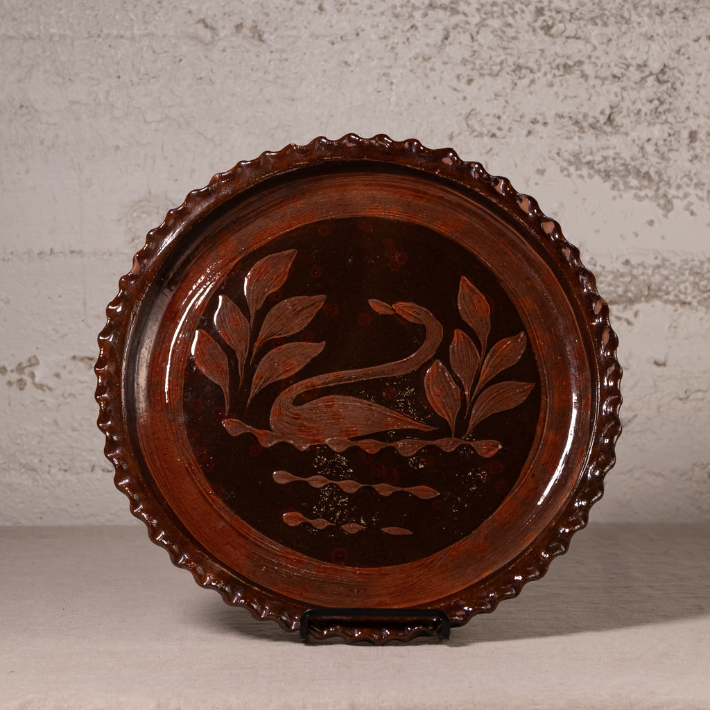 A round dark brown wooden serving tray with a scalloped edge, featuring an inlaid swan and leaf design in the center, displayed on a stand against a textured light gray wall.
