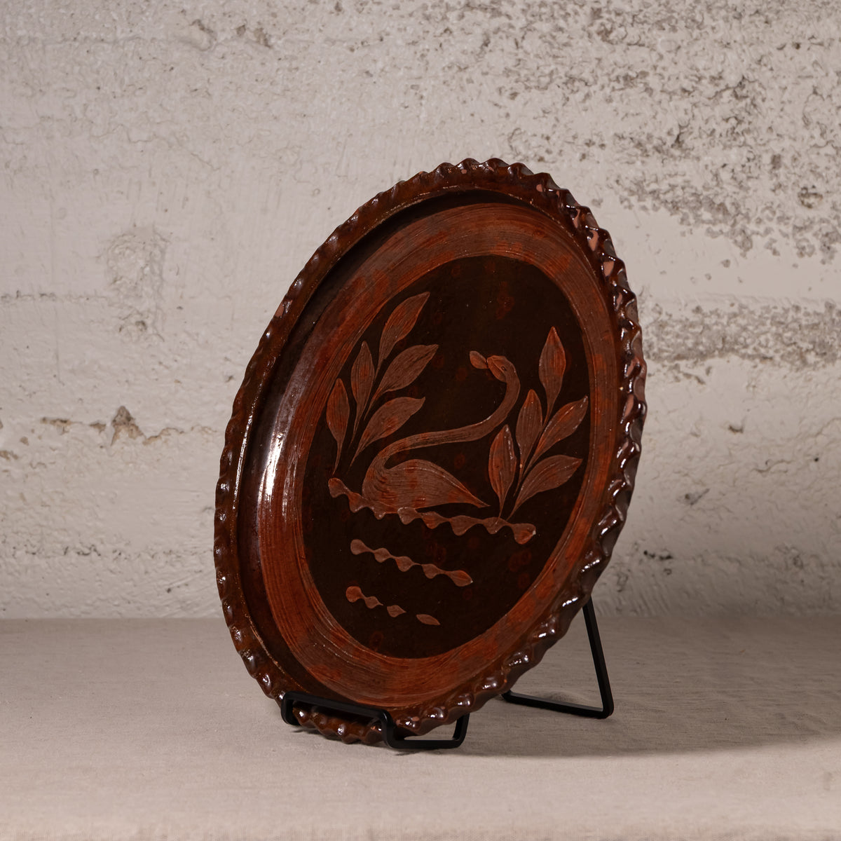An oval dark brown wooden decorative tray with a carved swan and leaf design in the center, displayed on a black metal stand against a textured light gray wall.