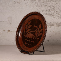 An oval dark brown wooden decorative tray with a carved swan and leaf design in the center, displayed on a black metal stand against a textured light gray wall.