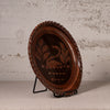 An oval dark brown wooden decorative tray with a swan and leaf design inlaid in the center, displayed on a black metal stand against a textured light gray wall.