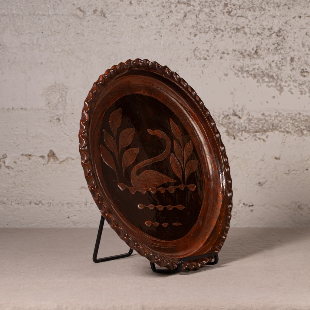 An oval dark brown wooden decorative tray with a swan and leaf design inlaid in the center, displayed on a black metal stand against a textured light gray wall.