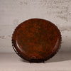 A round, scalloped-edge decorative plate or tray with a glossy dark brown glaze and orange floral patterns, displayed on a small black stand against a textured light gray wall.