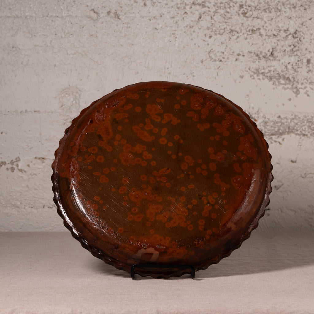 A round, scalloped-edge decorative plate or tray with a glossy dark brown glaze and orange floral patterns, displayed on a small black stand against a textured light gray wall.