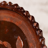Close-up detail of a dark brown ceramic plate with a glossy finish, showing a scalloped rim and a leaf motif, set against a light neutral background.
