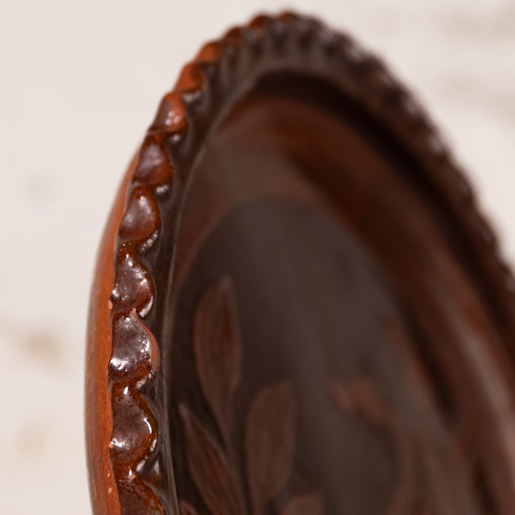 Close-up of a dark brown carved wooden tray or plate with a scalloped rim and leaf-like relief details, photographed against a softly blurred light background.